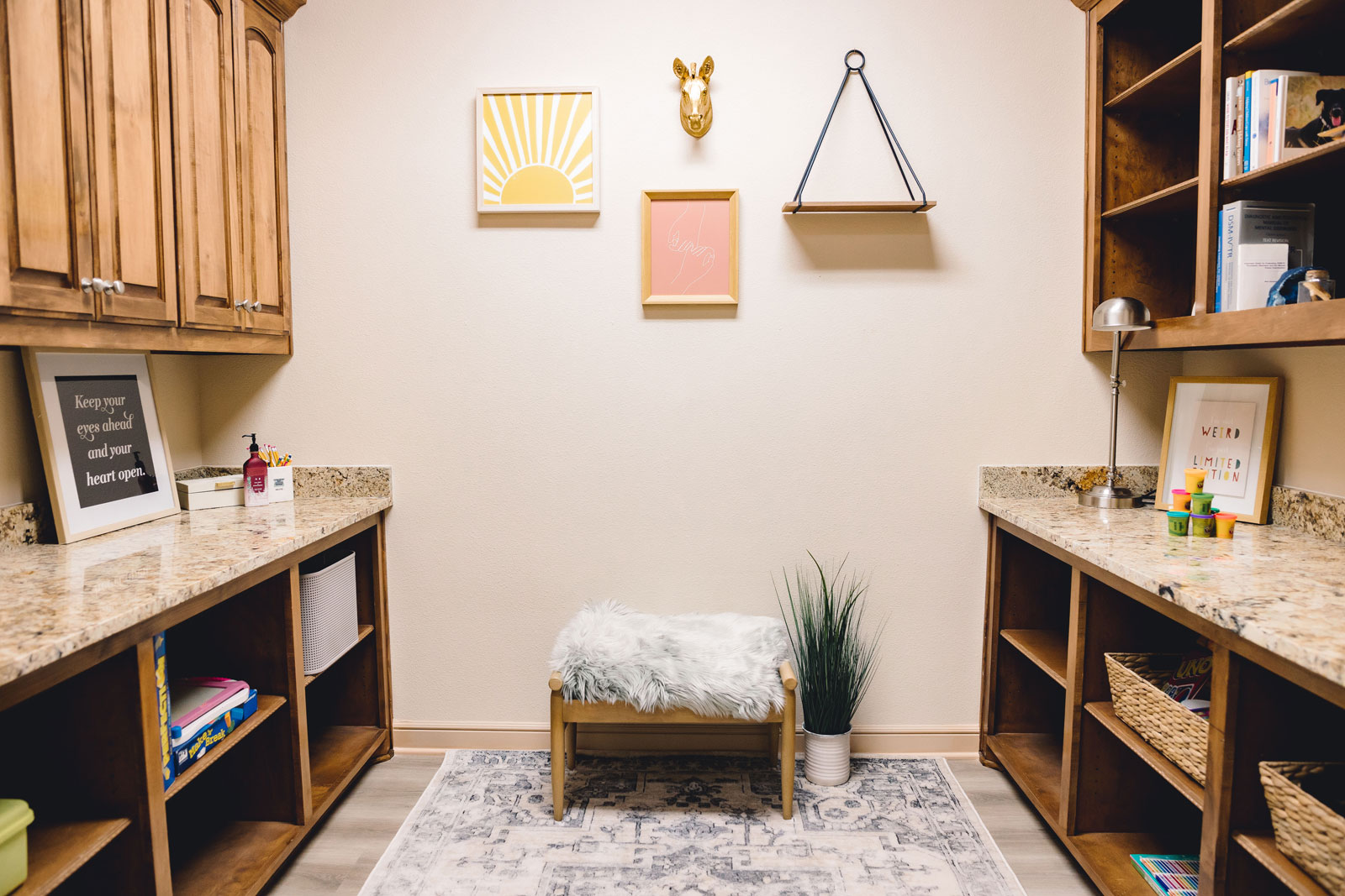 Cozy office or playroom with wooden cabinets, granite countertops, wall art, a soft bench, and decorative plants